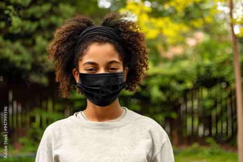 Teenage girl smiling behing stylish protective mask. Beautiful teenage girl smiling behind her stylish protective black mask. She his mixed-race and is looking at the camera. Nature in background. Hor photo
