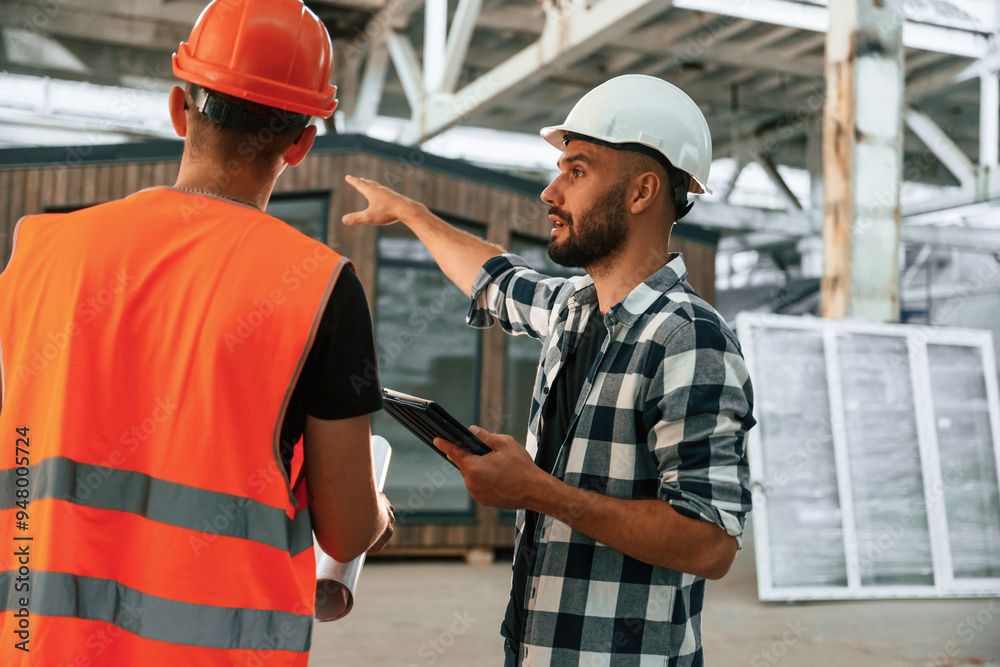 Builder is talking with manager or architect. Two workers are in the industrial wooden warehouse