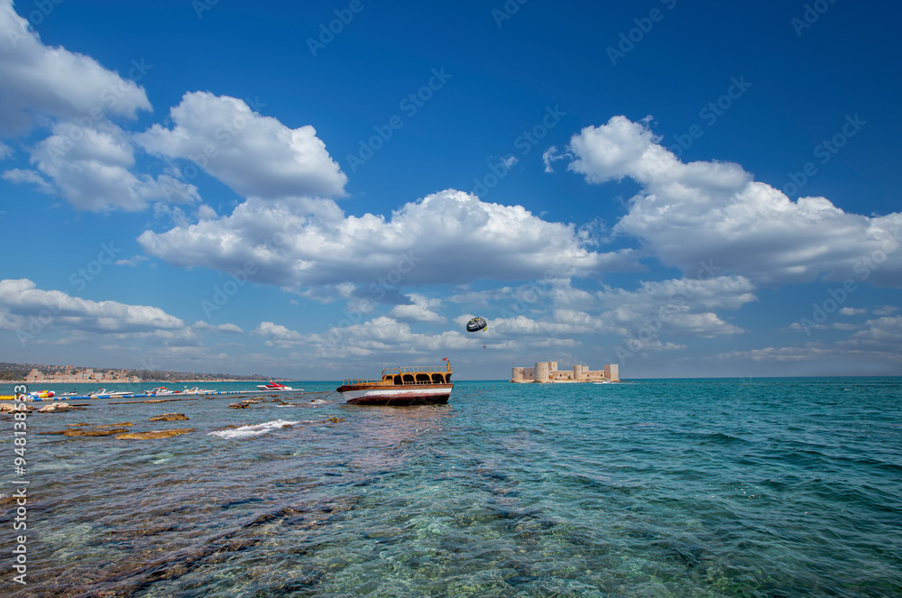 Maiden Castle, Korykos Castle in the background, a boat resting on the rocks on the beach in the foreground and a speedboat flying people with parachutes - Mersin, Turkey.