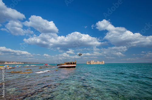 Obraz na płótnie Maiden Castle, Korykos Castle in the background, a boat resting on the rocks on the beach in the foreground and a speedboat flying people with parachutes - Mersin, Turkey. w salonie