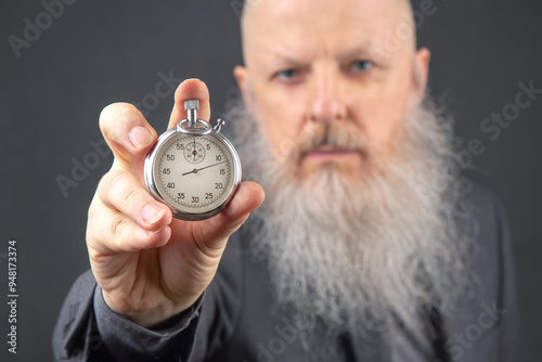 experienced man with a substantial gray beard demonstrating the time on a mechanical stopwatch, positioned in front of a dark gray background, illustrating meticulousness and time awareness photo