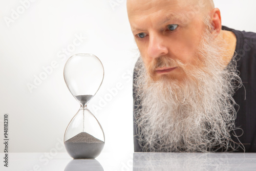 An adult man with a large white beard looks at the falling grains of sand in an hourglass with regret and anxiety. the concept of the transience of time. passage of time. photo