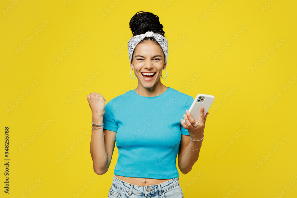 Young happy Latin woman she wears blue top t-shirt headband casual clothes hold in hand use mobile cell phone do winner gesture isolated on plain yellow background studio portrait. Lifestyle concept. Young happy Latin woman she wears blue top t-shirt headband casual clothes hold in hand use mobile cell phone do winner gesture isolated on plain yellow background studio portrait. Lifestyle concept.