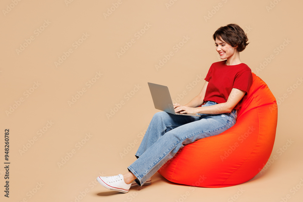 Full body young happy IT woman wear red t-shirt casual clothes sit in bag chair hold use work on laptop pc computer isolated on plain pastel light beige background studio portrait. Lifestyle concept.