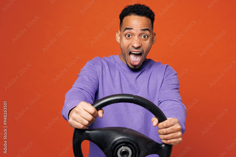 Young scared fearful shocked man of African American ethnicity wear purple sweatshirt casual clothes hold steering wheel driving car isolated on plain red orange background studio. Lifestyle concept. Young scared fearful shocked man of African American ethnicity wear purple sweatshirt casual clothes hold steering wheel driving car isolated on plain red orange background studio. Lifestyle concept.
