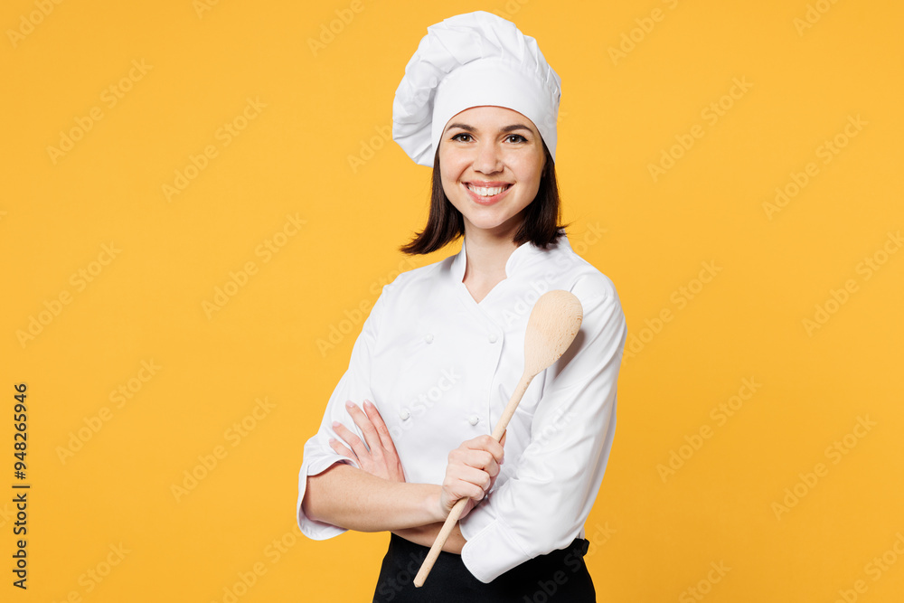 Young smiling happy cheerful fun chef cook baker woman wear white shirt black apron uniform toque chefs hat hold in hand wooden spoon isolated on plain yellow background studio. Cooking food concept. Young smiling happy cheerful fun chef cook baker woman wear white shirt black apron uniform toque chefs hat hold in hand wooden spoon isolated on plain yellow background studio. Cooking food concept.