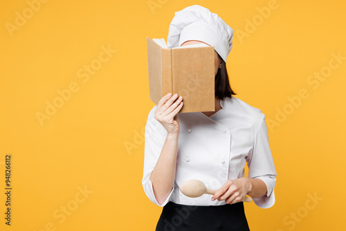 Young chef cook baker woman she wear white shirt black apron uniform toque chefs hat hold in hand cover face with book reading recipe isolated on plain yellow background studio. Cooking food concept. photo