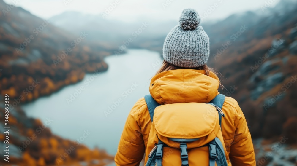 A person wearing a yellow jacket and grey woolen hat with a backpack, standing facing a serene lake surrounded by mountains and autumn foliage, capturing tranquility and adventure. A person wearing a yellow jacket and grey woolen hat with a backpack, standing facing a serene lake surrounded by mountains and autumn foliage, capturing tranquility and adventure.