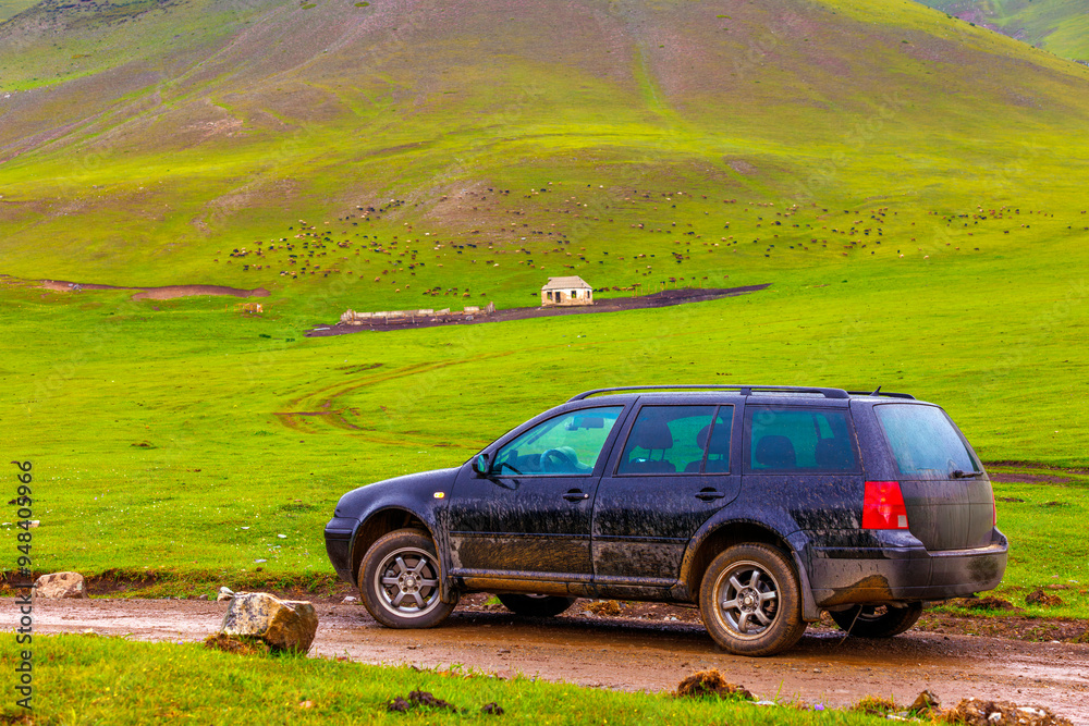 black car on a dirt road in the mountain hills with distant flock of sheep grazing on green pasture at rainy day near farm ruins black car on a dirt road in the mountain hills with distant flock of sheep grazing on green pasture at rainy day near farm ruins