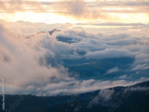 Majestic clouds envelop the rugged mountain peaks as the sun sets, casting a warm glow across the valley