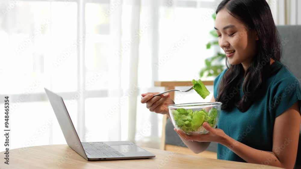 Young asian woman enjoying a healthy salad while working remotely at her laptop Young asian woman enjoying a healthy salad while working remotely at her laptop