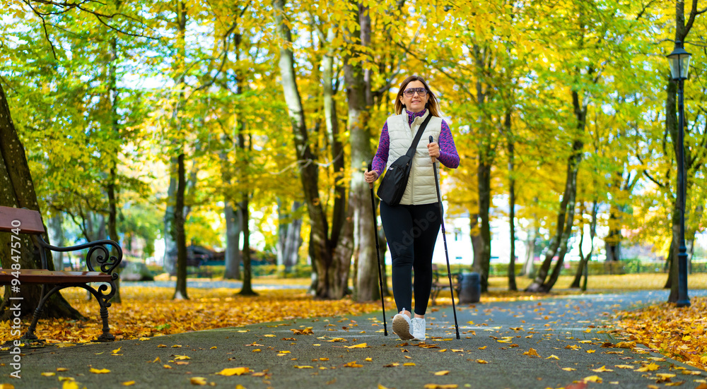 Fototapeta premium Beautiful middle-aged blonde woman dressed sporty exercising nordic walking on alley full of yellow fallen leaves in park on autumn day. Front view. Autumnal training of Nordic walking.