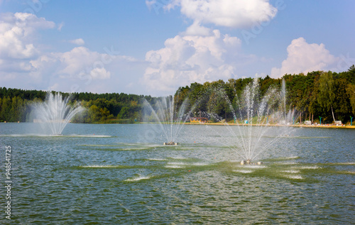 Fototapeta Naklejka Na Ścianę i Meble -  Lake with water cannon and fountain breeze, green leafy trees in the background, sunny summer day with clouds in Europe