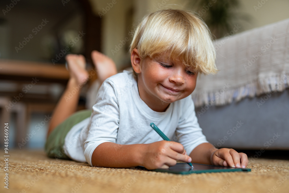 Blonde Boy Lying on the Floor Drawing on a Tablet with a Pen Blonde Boy Lying on the Floor Drawing on a Tablet with a Pen