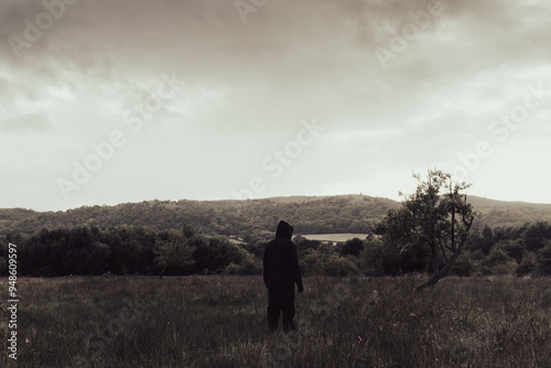 A strange hooded figure, back to camera. Standing in a field in the countryside. On a moody stormy cloudy day