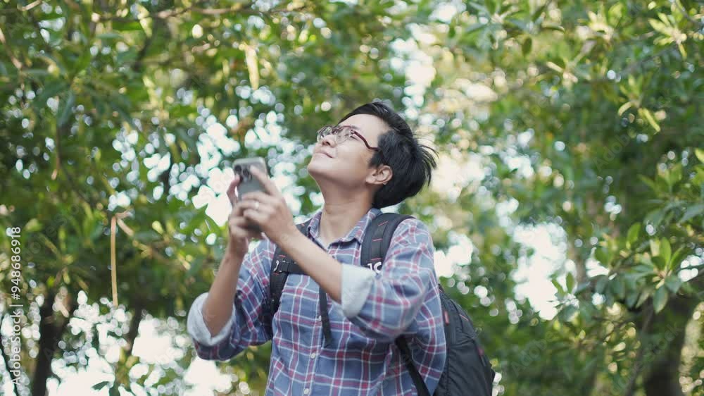 A young, confident Asian woman with short hair is walking through a lush forest with taking photo from her smartphone. She is enjoying the serenity and greenery with freedom of the nature outdoors. A young, confident Asian woman with short hair is walking through a lush forest with taking photo from her smartphone. She is enjoying the serenity and greenery with freedom of the nature outdoors.