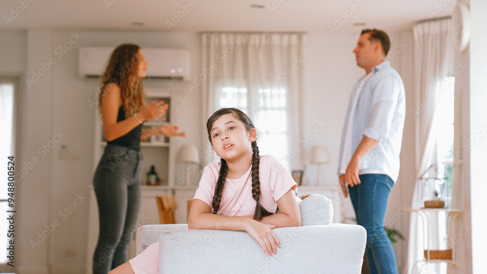 Fototapeta premium Annoyed and unhappy young girl sitting on sofa trapped in middle of tension by her parent argument in living room. Unhealthy domestic lifestyle and traumatic childhood develop to depression Synchronos