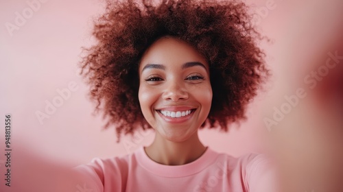 An exuberant woman with afro hair takes a close-up selfie wearing a pink outfit, radiating happiness and confidence against a pink backdrop in a vibrant, modern style.