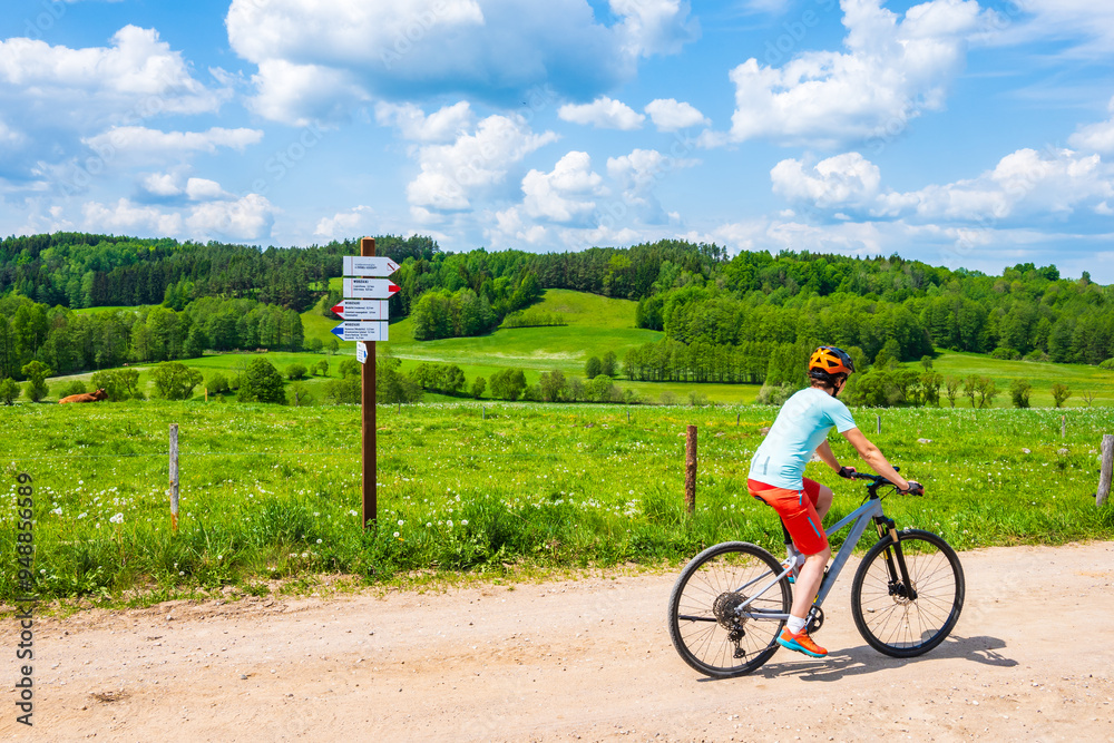 Woman cyclist riding bicycle on gravel rural road among green meadows, Suwalski Landscape Park, Podlasie, Poland