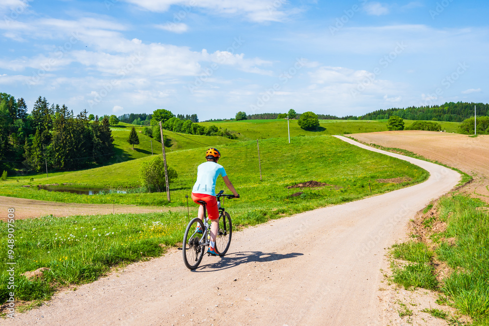 Obraz premium Woman cyclist riding bicycle on gravel rural road among green meadows and fields, Suwalski Landscape Park, Podlasie, Poland