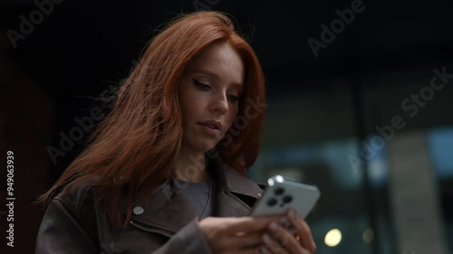 Low-angle view of cheerful cute young woman wearing fashion leather jacket using typing mobile phone smiling looking to screen, happily using smartphone app walking in city on cloudy day.