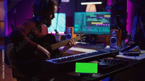 Songwriter playing acoustic guitar in home studio with greenscreen display running on smartphone app. Musician practices singing on musical instrument with strings, isolated screen. Camera B.