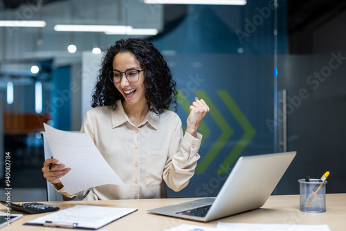 Happy businesswoman celebrating achievement in office while reviewing important documents. Woman seated at desk with laptop expressing excitement and success. Positive and enthusiastic office moment.