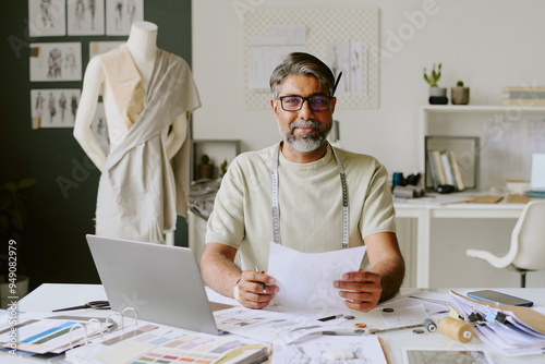 Portrait of excited tailor with measurement tape on shoulders holding fresh sketch and looking at camera photo