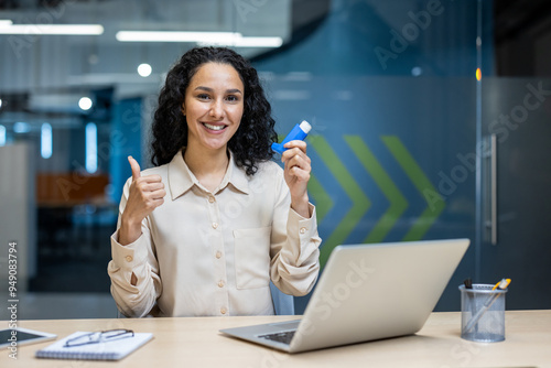 Wallpaper Mural Confident woman in professional office attire holding asthma inhaler while showing a thumbs up sign. She sits at a desk with laptop and notes, conveying health and positivity in workplace setting Torontodigital.ca
