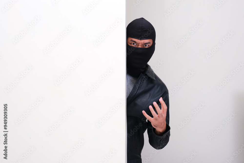 A thief wearing balaclava peeking behind wall showing fierce expression A thief wearing balaclava peeking behind wall showing fierce expression