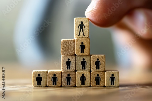 Building a Strong Team: A hand carefully positions a wooden block with a human figure atop a pyramid of similar blocks, illustrating the concept of leadership, teamwork, and organizational structure.  photo