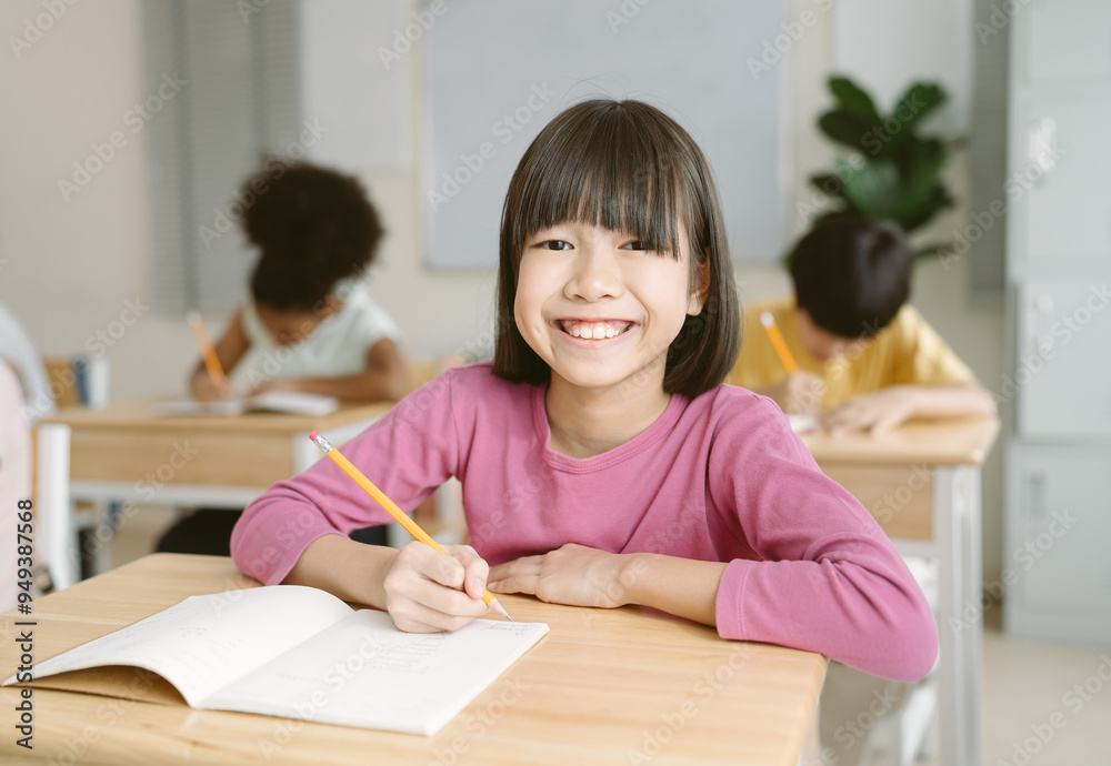 Portrait of Smile Asian schoolgirl write on notebook while sitting on desk in classroom and looking at camera at primary school. Education, elementary school, learning concept. Portrait of Smile Asian schoolgirl write on notebook while sitting on desk in classroom and looking at camera at primary school. Education, elementary school, learning concept.