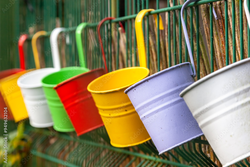 A row of coloured metal buckets hanging from a fence in an outdoor setting A row of coloured metal buckets hanging from a fence in an outdoor setting