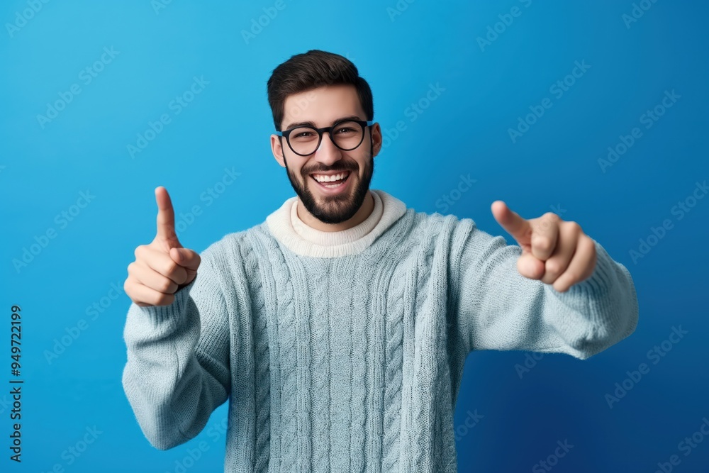 Young handsome man with beard wears casual sweater, glasses over blue background. Points to camera with fingers, smiling positive, cheerful. Attractive male fashion model with confident, friendly Young handsome man with beard wears casual sweater, glasses over blue background. Points to camera with fingers, smiling positive, cheerful. Attractive male fashion model with confident, friendly