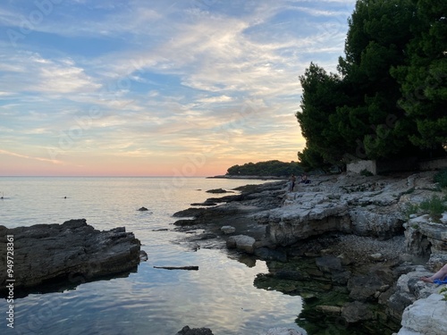 Sunset in Croatia, Pula. Amazing view of the orange sunset with blue sky and clouds on the sea and some trees in the forest in the background with rocky beach. 