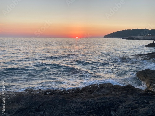 Sunset in Croatia, Pula. Amazing view of the orange sunset with blue sky and clouds on the sea and some trees in the forest in the background with rocky beach. 