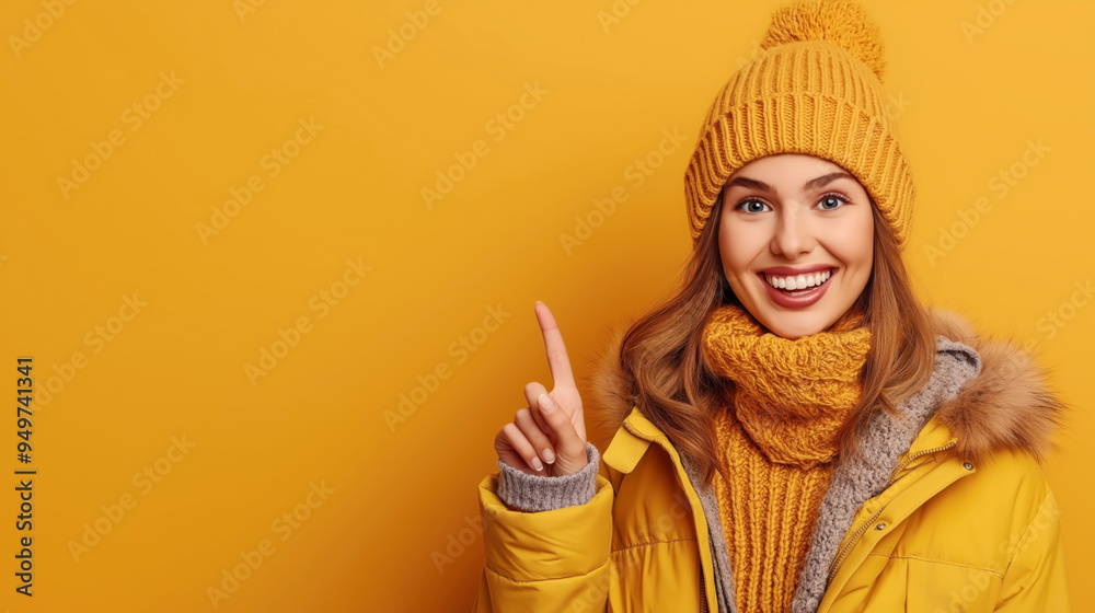 Smiling woman wearing a yellow knit hat, scarf, and winter jacket with a fur hood, pointing upwards against matching yellow background. Smiling woman wearing a yellow knit hat, scarf, and winter jacket with a fur hood, pointing upwards against matching yellow background.