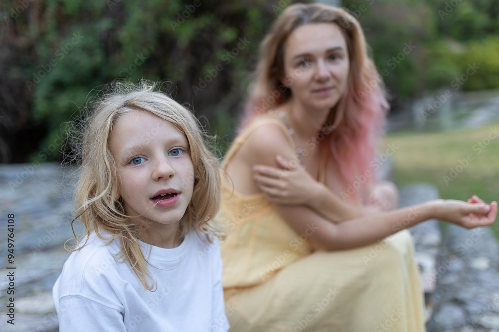 Young blonde girl looking pensive while sitting with her mother outdoors Young blonde girl looking pensive while sitting with her mother outdoors