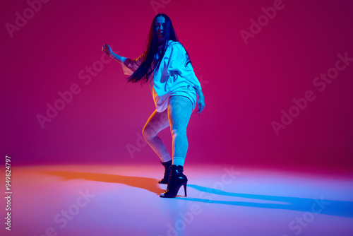 Artistic, talented, beautiful young woman in oversized clothes and heeled shoes dancing high heel dance against magenta studio background. Concept of hobby, entertainment, performance, creativity