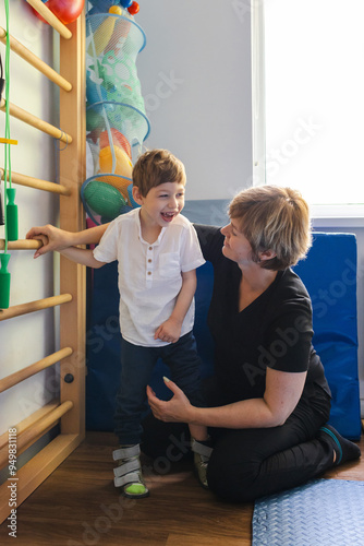 Young boy with cerebral palsy in orthopedic shoes for fine standing and walking happily participates in rehabilitation exercises with physio therapist in rehab clinic photo