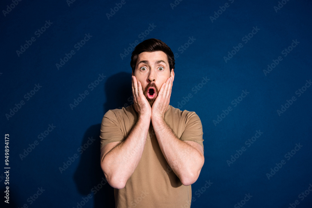 Photo of speechless man with beard dressed beige t-shirt astonished staring hold hands on cheeks isolated on dark blue color background Photo of speechless man with beard dressed beige t-shirt astonished staring hold hands on cheeks isolated on dark blue color background