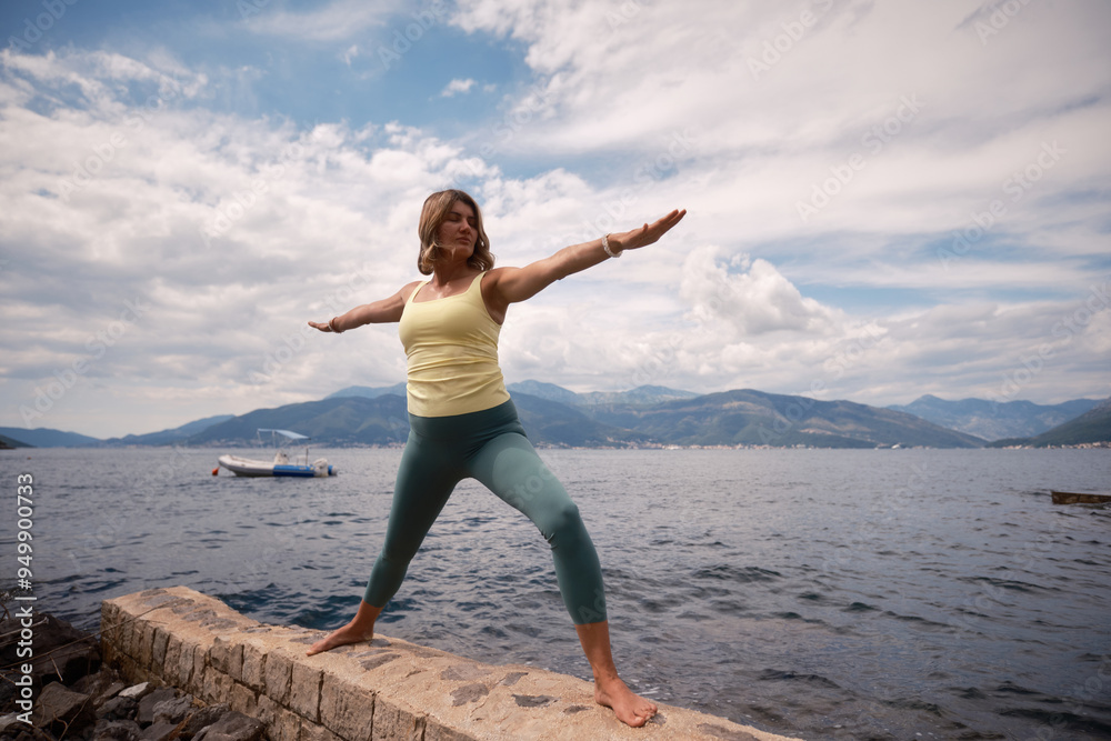 Obraz premium A woman practicing yoga in warrior pose by the serene lake under a cloudy sky