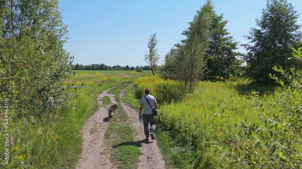A man in light clothing and linen pants walks his dog through wild fields and trees, enjoying a sunny, clear day with a bag over his shoulder. A man in light clothing and linen pants walks his dog through wild fields and trees, enjoying a sunny, clear day with a bag over his shoulder.