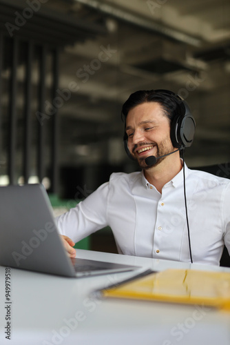 A man wearing a headset is smiling while using a laptop. He is likely working on a project or communicating with someone. Concept of productivity and focus