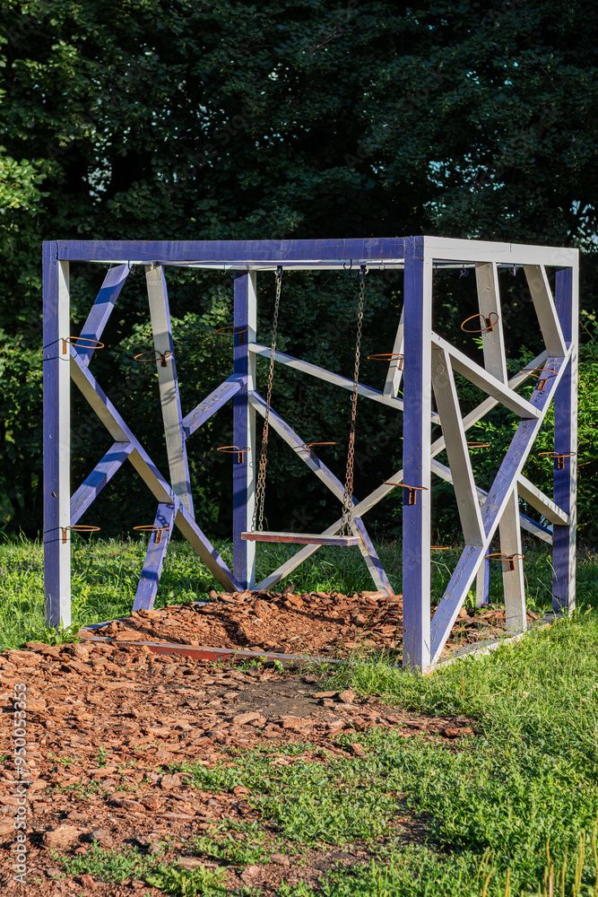 Abstract geometric structure made of wood with swings in a park, surrounded by greenery and trees in bright sunlight.
Abstract geometric structure made of wood with swings in a park, surrounded by greenery and trees in bright sunlight.