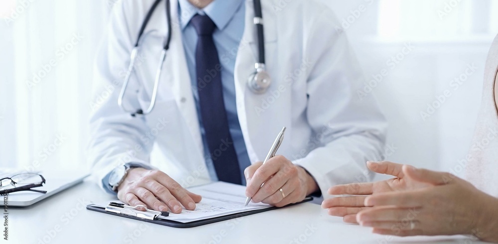 A doctor and a patient. The physician, wearing a white medical coat over a blue shirt and tie, is filling out a medical record form during a consultation in the clinic. Medical service A doctor and a patient. The physician, wearing a white medical coat over a blue shirt and tie, is filling out a medical record form during a consultation in the clinic. Medical service