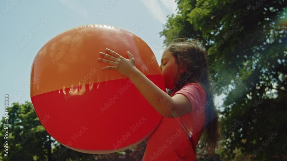 A cheerful girl works on inflating a big beach ball in the park, taking advantage of the beautiful summer weather. The scene captures the simple joys of outdoor playtime.