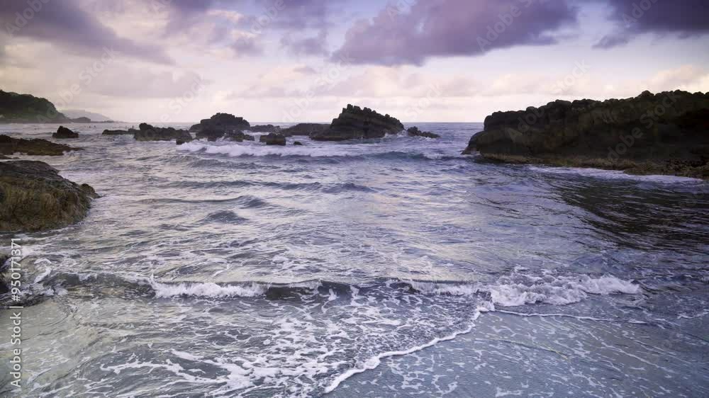 At dusk. Gentle waves lap against smooth, rounded rocks in a tranquil cove. The sky is painted in soft hues of pink and purple, reflecting on the calm water. Suao, Yilan, Taiwan. At dusk. Gentle waves lap against smooth, rounded rocks in a tranquil cove. The sky is painted in soft hues of pink and purple, reflecting on the calm water. Suao, Yilan, Taiwan.