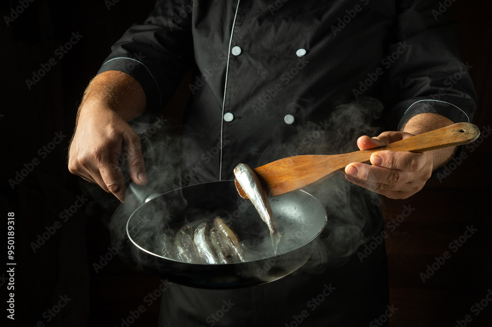 The chef fries capelin fish in a frying pan. The spatula in the cook hand for turning the fish. The concept of a fish diet The chef fries capelin fish in a frying pan. The spatula in the cook hand for turning the fish. The concept of a fish diet