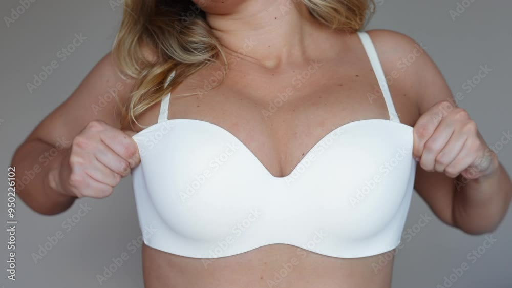 Young blonde woman pulling her bra up lifting her saggy breasts on a grey background. The result of breastfeeding